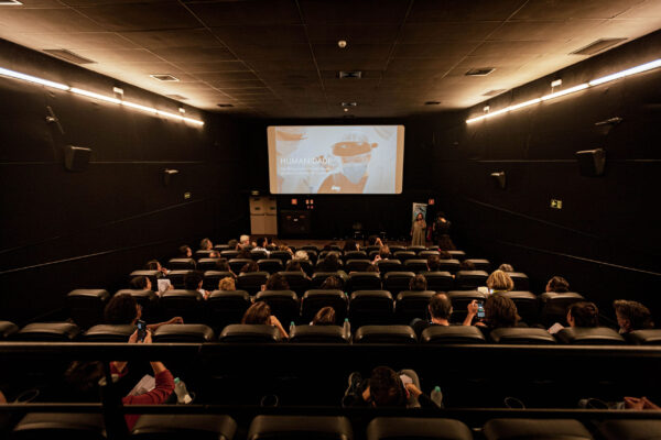 Em um cinema estão diversas pessoas sentadas vendo uma tela com o banner do curta-metragem Humanidade. Está escuro.